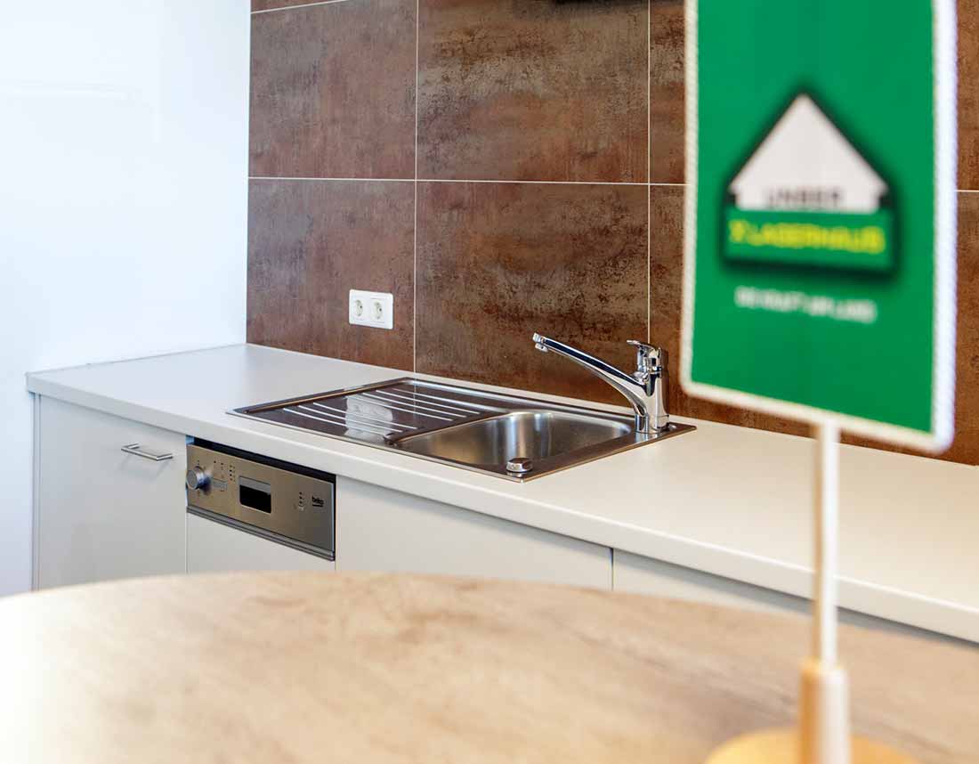 Büro-Teeküche mit Edelstahlspüle, Geschirrspüler und brauner Fliesenwand, im Vordergrund ein grünes Schild.