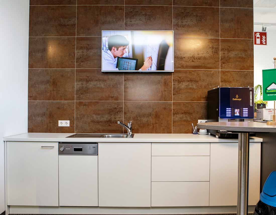 Büro-Teeküche mit Spüle, Geschirrspüler, Kaffeemaschine, braunen Fliesen und an der Wand montiertem Fernseher.