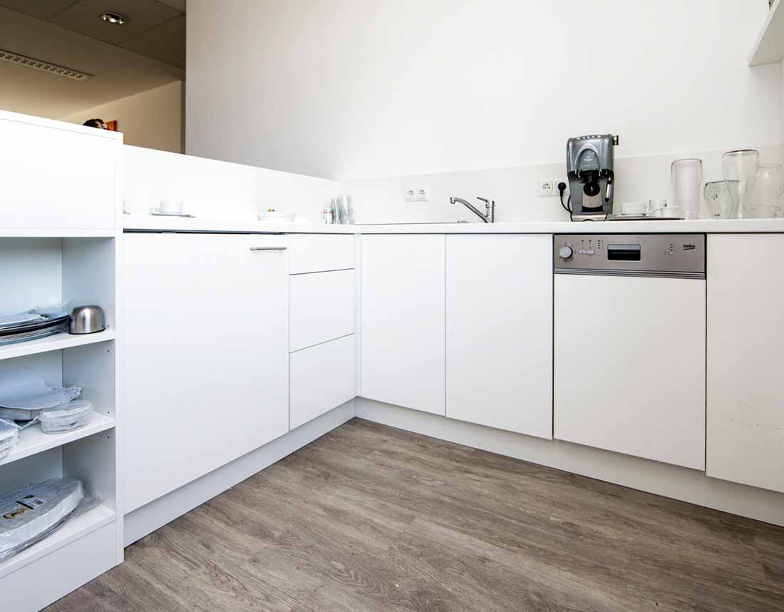 Helle Büro-Teeküche mit weißer Einrichtung, Geschirrspüler, Kaffeemaschine und Regalen.
