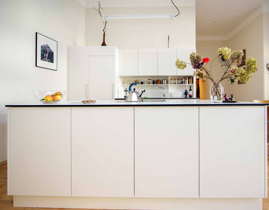 Moderne, weiße Küche mit Insel, dekoriert mit Obstschale und Blumen, im offenen Wohnraum.