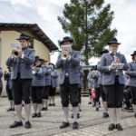 Eine Blaskapelle in traditioneller Tracht spielt auf einem Platz, die Musiker tragen Instrumente und Notenständer.
