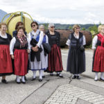 Eine Gruppe von Frauen in traditioneller Tracht steht zusammen mit einem Mann im Anzug auf einer Straße.