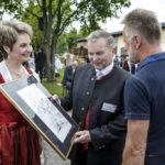 Ein Mann überreicht ein eingerahmtes Bild an eine Frau in rotem Dirndl, während ein anderer Mann zusieht.