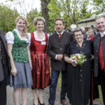 Sechs Personen in traditioneller Tracht posieren bei einem Fest im Freien, eine Frau hält Blumen.