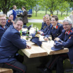 Feuerwehrmänner in blauen Uniformen sitzen lächelnd an einem Holztisch und genießen das Fest im Freien.