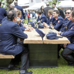 Mehrere Feuerwehrmänner in Uniformen sitzen an einem langen Holztisch und unterhalten sich bei einem Fest.