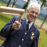 Ein lachender älterer Mann in Uniform hält ein handgefertigtes Holzobjekt beim Jubiläumsfest.