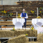 Ein Mann in traditioneller Kleidung hält eine Rede auf einer Bühne, dekoriert mit Blumen und Stroh.