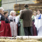 Frauen in roten und grünen Trachten singen, dirigiert von einem Mann in braunem Mantel.
