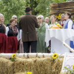 Frauen in traditionellen Trachten singen unter der Leitung eines Mannes auf einer Bühne aus Heuballen.