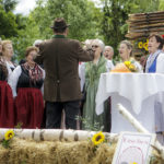 Eine Gruppe von Frauen in Trachten singt unter der Leitung eines Mannes auf einer Bühne mit Heuballen.