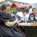 Ältere Frau sitzt an einem Tisch, im Hintergrund Feuerwehrleute in Uniform, mehrere Weingläser auf dem Tisch.