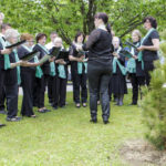 Ein Chor mit grünen Schals singt draußen unter Bäumen, dirigiert von einer Frau in Schwarz.