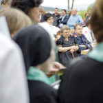 Ältere Frau sitzt mit Familie und Freunden, während vorne eine Veranstaltung stattfindet.