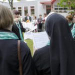 Eine Gruppe von Frauen, teils in Chorkleidung, singt zusammen, während die Chorleiterin dirigiert.