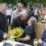 Ein Mann überreicht einer älteren Frau einen Blumenstrauß, während mehrere Personen freundlich zuschauen.