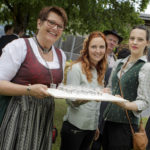 Drei Frauen, eine in Tracht, bieten Getränke auf einem Tablett an und lächeln in die Kamera.