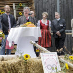 Ein Mann hält eine Rede am Mikrofon auf einer dekorierten Bühne, mehrere Personen stehen daneben.