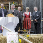 Ein Mann spricht ins Mikrofon auf einer festlich dekorierten Bühne, umgeben von mehreren Personen.