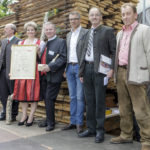 Sechs Personen stehen vor gestapeltem Holz, eine Frau hält eine große Ehrenurkunde.