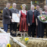 Eine Gruppe von Menschen posiert mit einer Ehrenurkunde und einem Blumenstrauß auf einer Bühne, umgeben von Strohballen und Sonnenblumen.