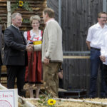 Ein Mann in traditioneller Kleidung erhält eine Auszeichnung und einen Handschlag auf einer dekorierten Bühne mit applaudierenden Personen im Hintergrund.
