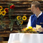 Mann in blauer Weste steht an einem dekorierten Tisch, hinter ihm Sonnenblumen vor einem Holzstapel.