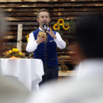 Ein Mann in blauer Weste spricht in ein Mikrofon, dekorierter Hintergrund mit Holzstapeln und Sonnenblumen.