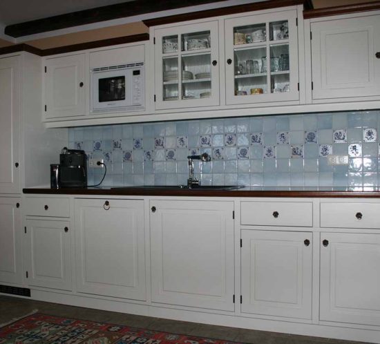 Weiße Massivholzküche im Rahmenbau mit Fliesenrückwand und integrierter Mikrowelle, Glasvitrine und Kaffeemaschine.