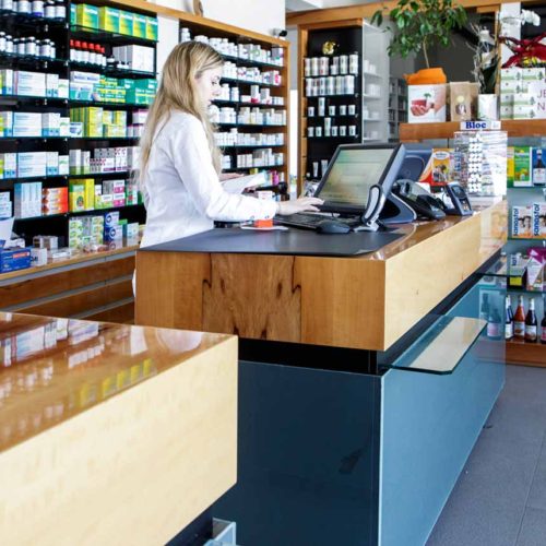 Apothekerin arbeitet an moderner Holztheke mit Medikamentregalen im Hintergrund.