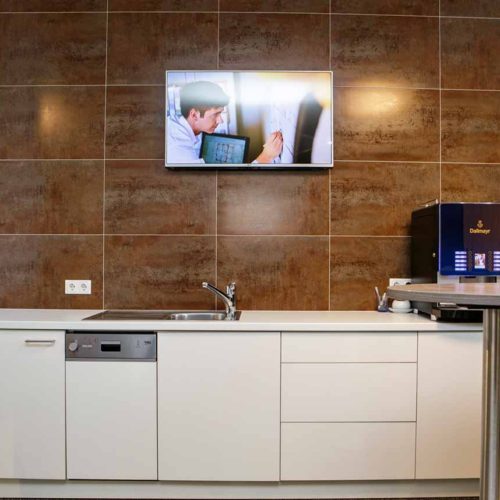 Büro-Teeküche mit Spüle, Geschirrspüler, Kaffeemaschine, braunen Fliesen und an der Wand montiertem Fernseher.