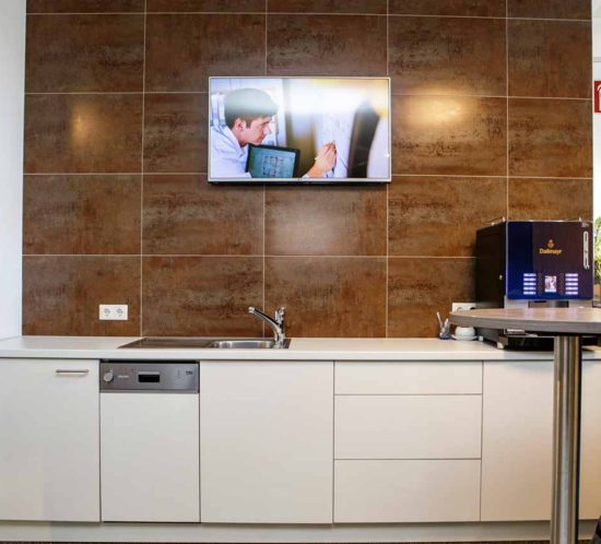 Büro-Teeküche mit Spüle, Geschirrspüler, Kaffeemaschine, braunen Fliesen und an der Wand montiertem Fernseher.