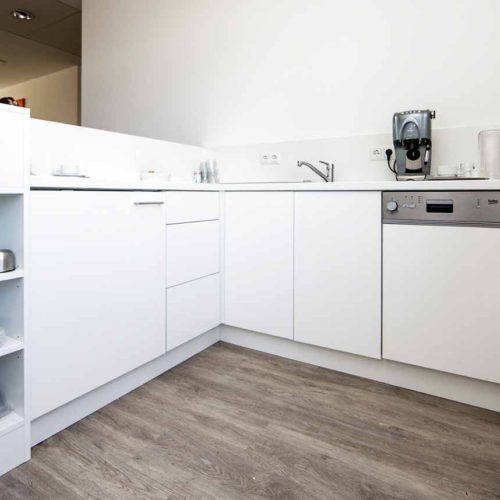 Helle Büro-Teeküche mit weißer Einrichtung, Geschirrspüler, Kaffeemaschine und Regalen.