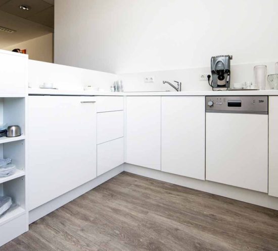 Helle Büro-Teeküche mit weißer Einrichtung, Geschirrspüler, Kaffeemaschine und Regalen.