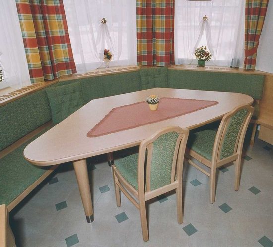 Eckbankgruppe mit grün gepolsterten Stühlen und Bank, hellem Holztisch, bunten Vorhängen und Tischdeko.