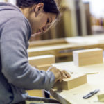 Eine Frau bearbeitet konzentriert ein kleines Holzstück in einer Werkstatt.