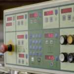 Nahaufnahme eines Steuerungspanels einer Maschine mit mehreren digitalen Anzeigen, Tasten und Not-Aus-Schalter.
