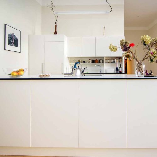 Moderne, weiße Küche mit Insel, dekoriert mit Obstschale und Blumen, im offenen Wohnraum.