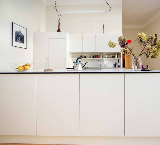 Moderne, weiße Küche mit Insel, dekoriert mit Obstschale und Blumen, im offenen Wohnraum.