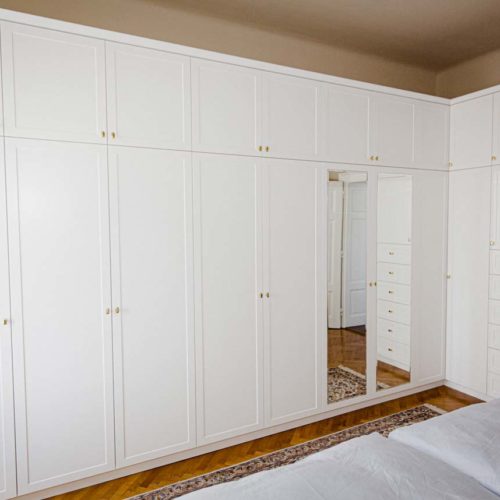 Großer weißer Kleiderschrank mit Spiegeln und goldenen Griffen, daneben mehrere Schubladen in einem Schlafzimmer.