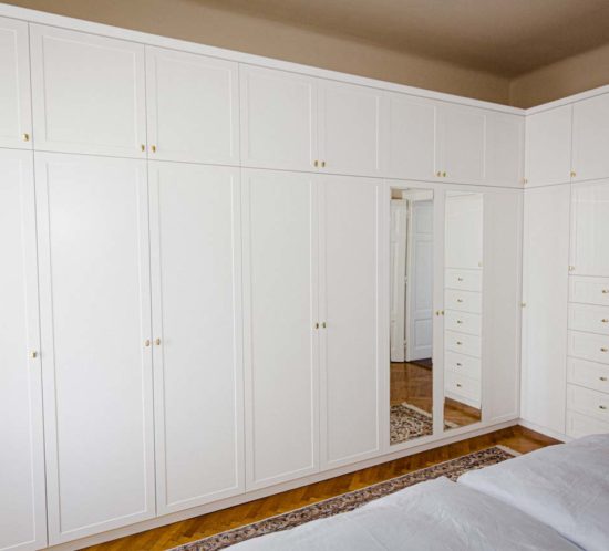 Großer weißer Kleiderschrank mit Spiegeln und goldenen Griffen, daneben mehrere Schubladen in einem Schlafzimmer.