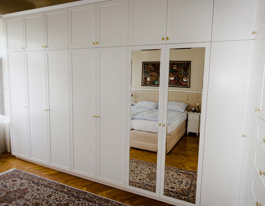 Großer weißer Kleiderschrank mit Spiegel und goldenen Griffen, reflektiert ein Bett im angrenzenden Raum.