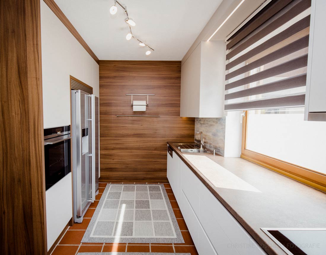 Moderne Küche mit Edelstahl-Kühlschrank, Holzverkleidung, großen Fenstern und gemustertem Teppich auf Terrakottafliesen.