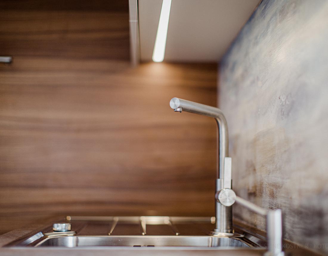 Nahaufnahme eines modernen Edelstahl-Wasserhahns in einer Küche mit Holzverkleidung und beleuchteter Rückwand.