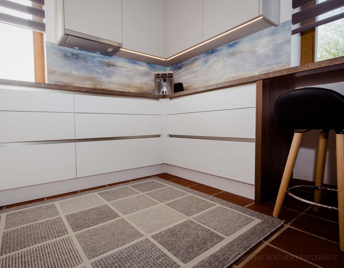Moderne Küche mit weißen Schubladen, gemusterter Rückwand, Barhocker und Teppich auf Terrakottafliesen.