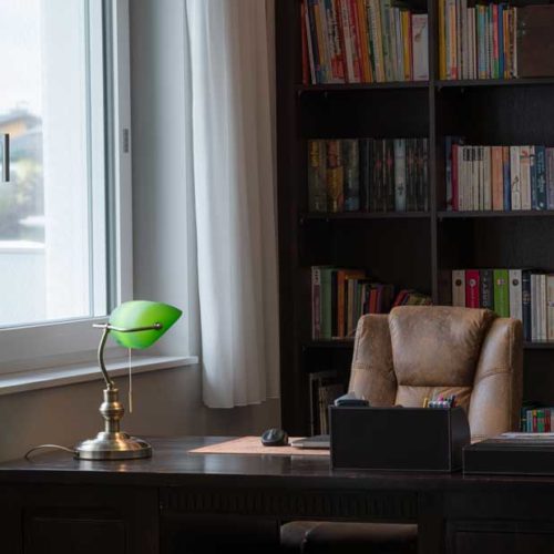 Büro mit großem Schreibtisch, Ledersessel, Bücherregal und grüner Schreibtischlampe neben einem Fenster.