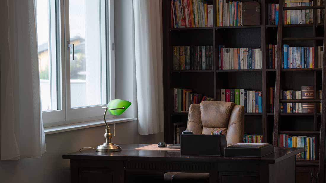 Büro mit großem Schreibtisch, Ledersessel, Bücherregal und grüner Schreibtischlampe neben einem Fenster.