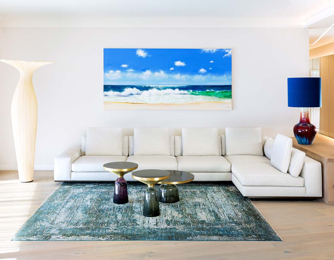 Modernes Wohnzimmer mit weißem Ecksofa, Küstenbild, bunten Beistelltischen und großen Stehlampen.