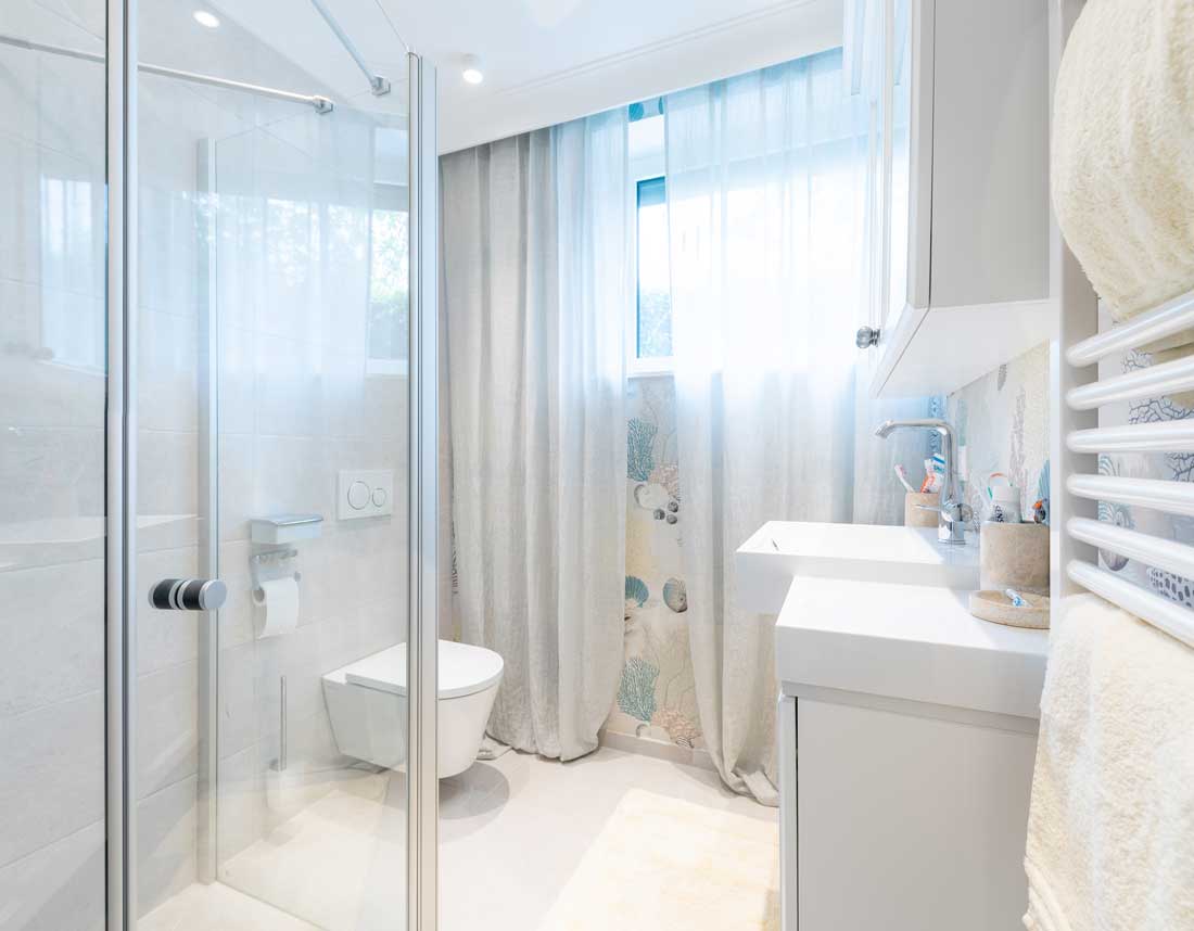 Helles Badezimmer mit Dusche, WC, Waschtisch und großen Vorhängen vor einem Fenster.
