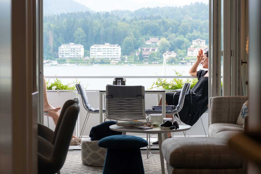 Zwei Personen sitzen auf einem Balkon mit Blick auf einen See und die umliegenden Berge.